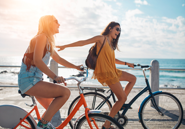 Zwei junge glückliche Frauen fahren an einem sonnigen Tag am Meer mit dem Fahrrad und haben gemeinsam Spass.