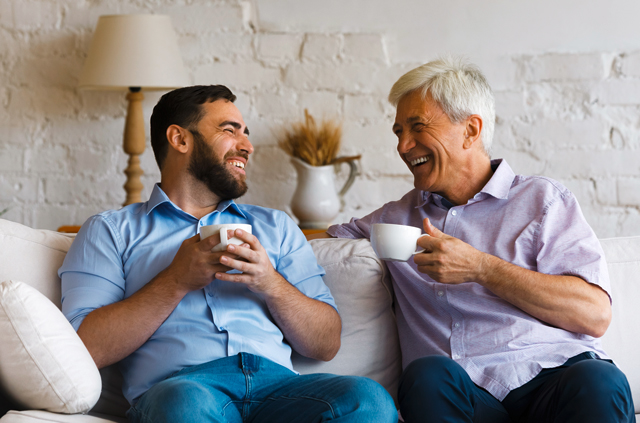 Zwei Maenner unterschiedlicher Generationen sitzen auf dem Sofa, trinken Kaffee und fuehren ein entspanntes und lustiges Gespraech.